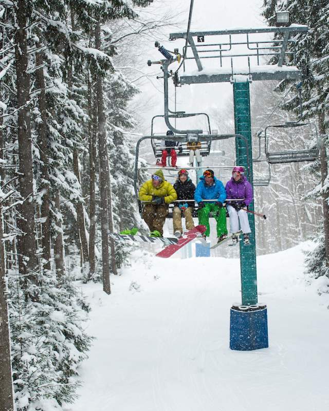 A group of four skiers in colorful outfits sits on a ski lift surrounded by snow-covered trees, conveying a joyful and adventurous winter scene.