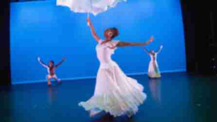 A trio of dancers dressed in white costumes perform on a stage with a blue background with the Alvin Ailey American Dance Theater