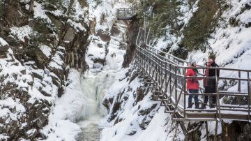 A snow-covered canyon with a frozen waterfall. Two people in winter clothing stand on a metal walkway, enjoying the serene, wintry view.