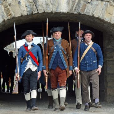 Three men dressed in historical military uniforms, holding muskets, walk under a stone arch. The scene conveys a sense of historical reenactment.